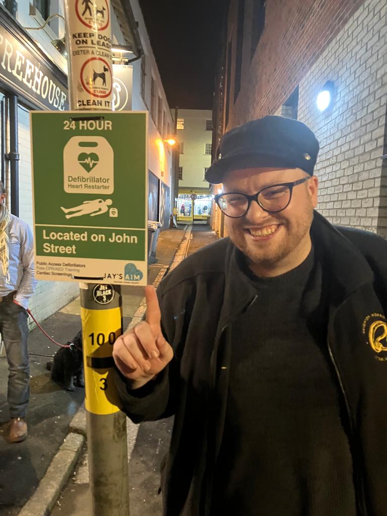 Jay, a Brighton Morris Musician, laughing as he finds his name on a lamp post sign for the nearest defibrillator at the the Inter-Varsity Folk Dance Festival, Exeter, February 2023