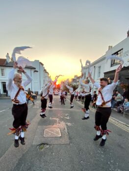 Six Brighton Morris dancers jumping high during a dance. The photo is taken along a street of terraced houses at sunset. the sun is in line with the street, so is framed by the dancers.